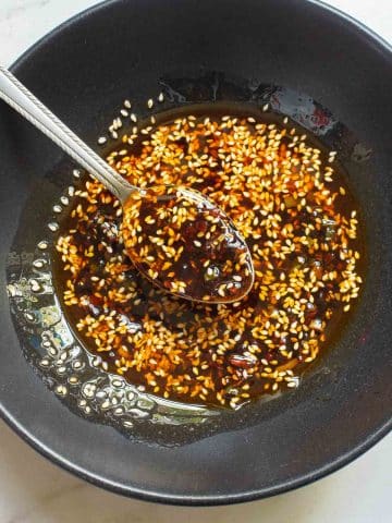 A black bowl brimming with a dark soy sauce mixture hints at garlic chili oil, its surface speckled with sesame seeds and chili flakes. A spoon rests within the aromatic blend, partially submerged. The bowl is elegantly placed on a light-colored surface, invitingly close to a plate of noodles.