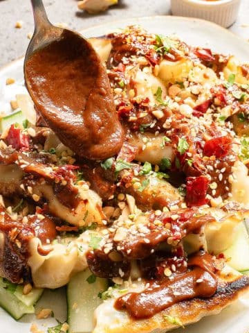 A spoon drizzles sauce over a plate of dumplings, garnished with chopped herbs, crushed nuts, and red pepper flakes. A small dish of chili oil is in the background on a textured surface.