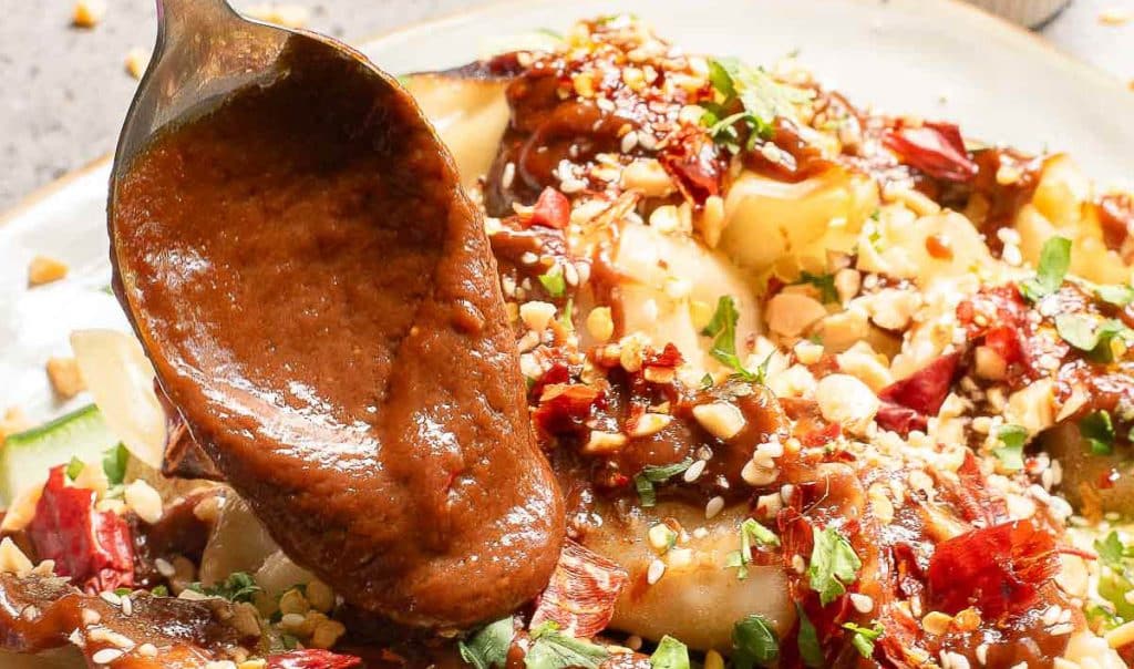 Close-up of a spoon pouring rich brown sauce over a dish topped with crispy red chili flakes, chopped peanuts, and fresh herbs on a white plate. The dish features thin noodles, suggesting a flavorful, spicy meal.