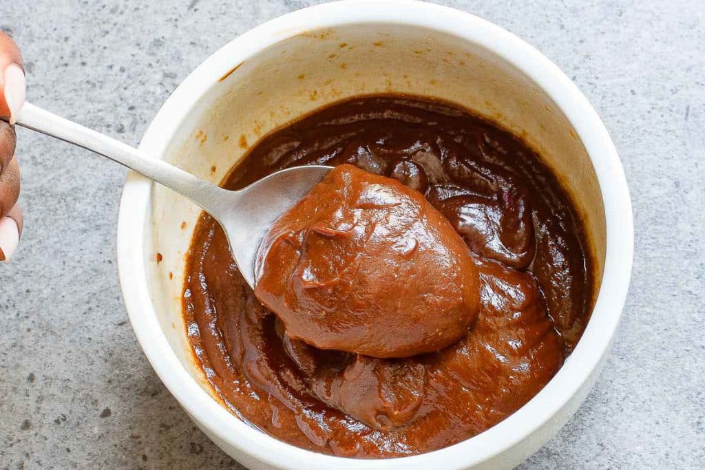A person holding a spoon filled with thick reddish-brown sauce above a white bowl containing more of the same sauce. The bowl is set on a gray textured surface.