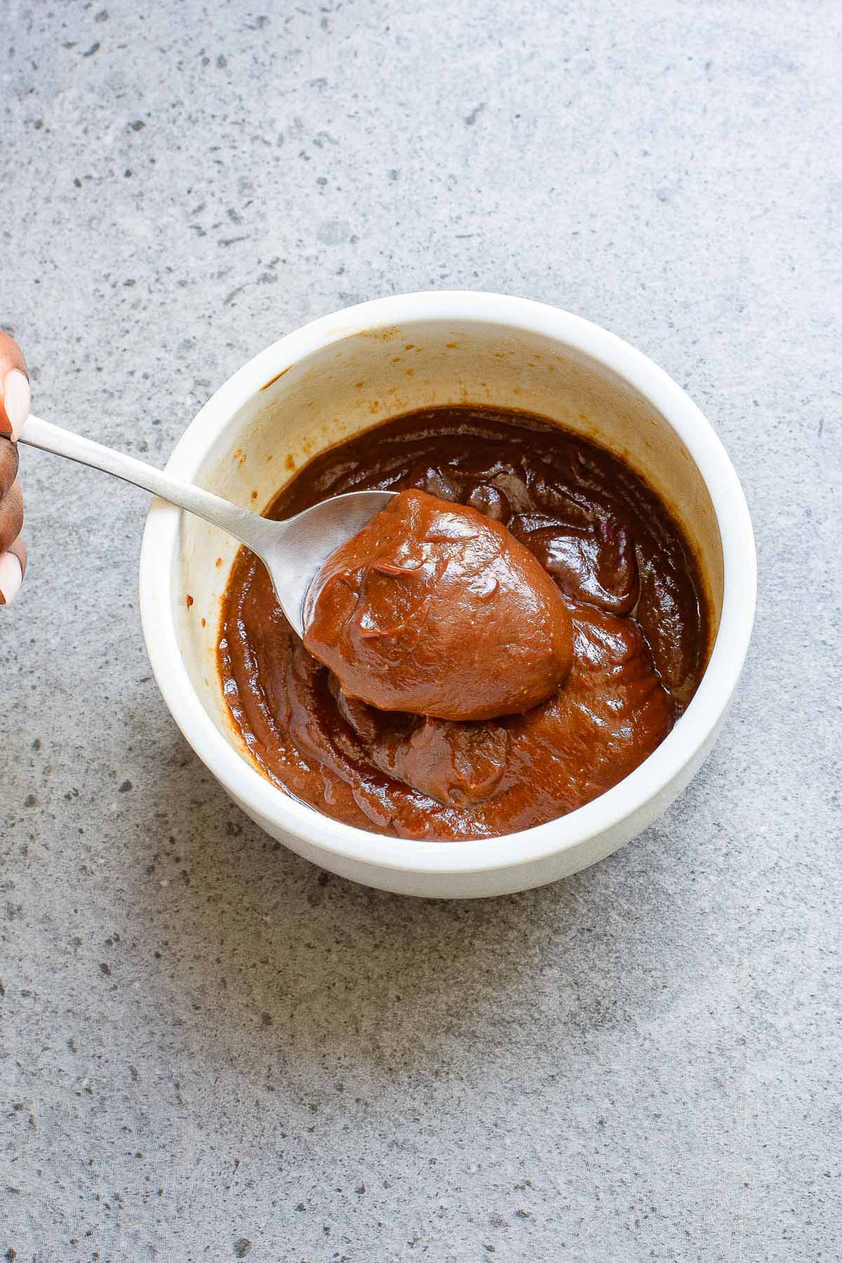 A person holds a spoon in a white bowl filled with thick, dark reddish-brown sauce on a speckled gray surface.