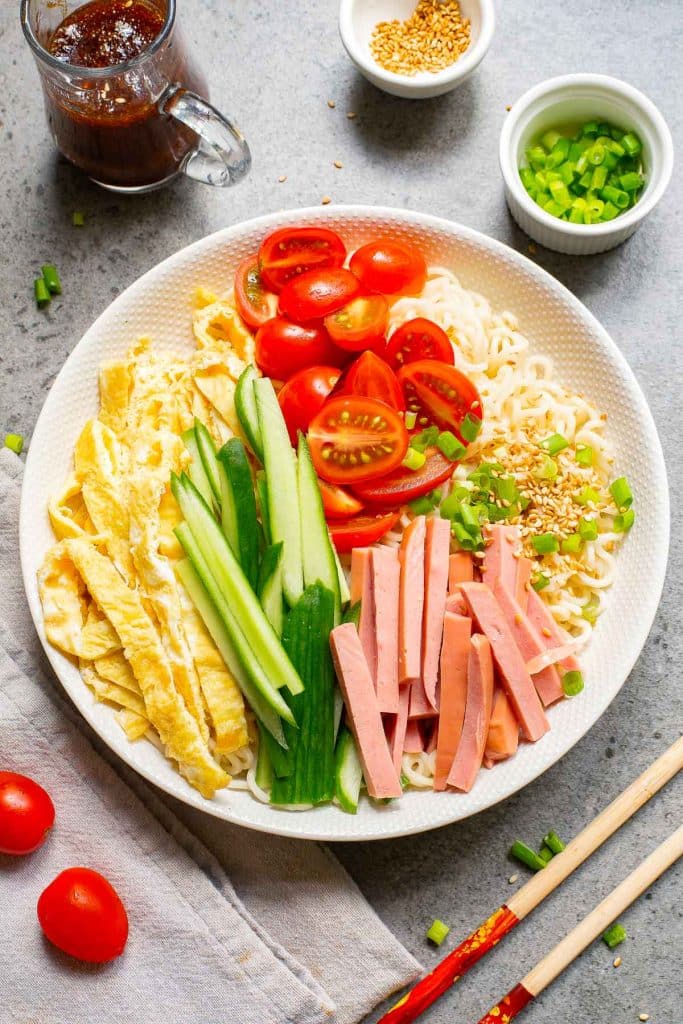 A round plate with cold noodles, sliced omelet, cucumber, ham, cherry tomatoes, and chopped green onions. A small bowl of sauce, sesame seeds, a dish with more onions, and a pair of chopsticks are nearby on a gray surface.