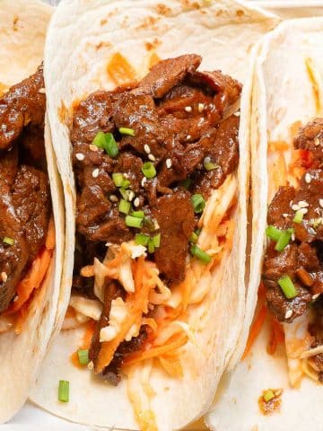 Three soft tacos filled with marinated beef, shredded carrots, and cabbage, garnished with chopped green onions and sesame seeds, displayed on a white plate against a gray background.