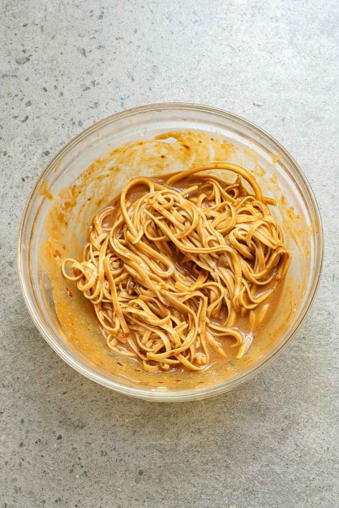 A clear glass bowl filled with noodles mixed in a creamy sauce, placed on a speckled gray surface. The sauce is thick and light brown, coating the noodles evenly.
