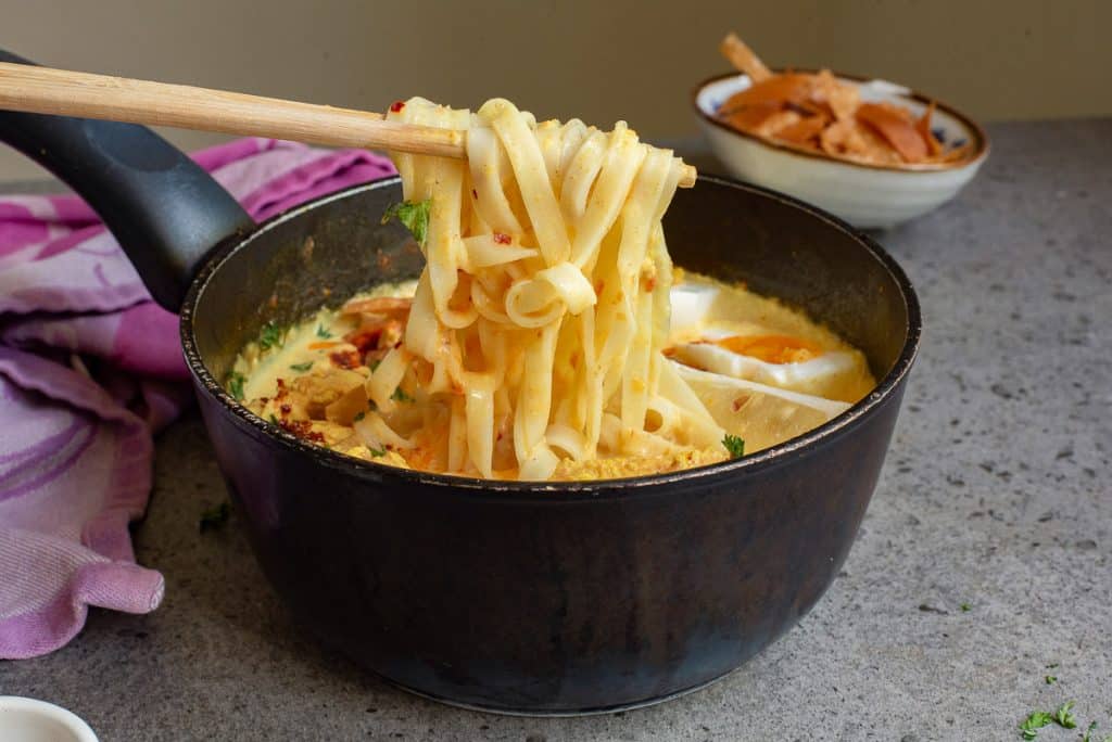 A black pot filled with creamy pasta, garnished with herbs, with a wooden fork lifting a portion. A pink cloth and a small bowl with bread are visible in the background, all on a gray countertop.