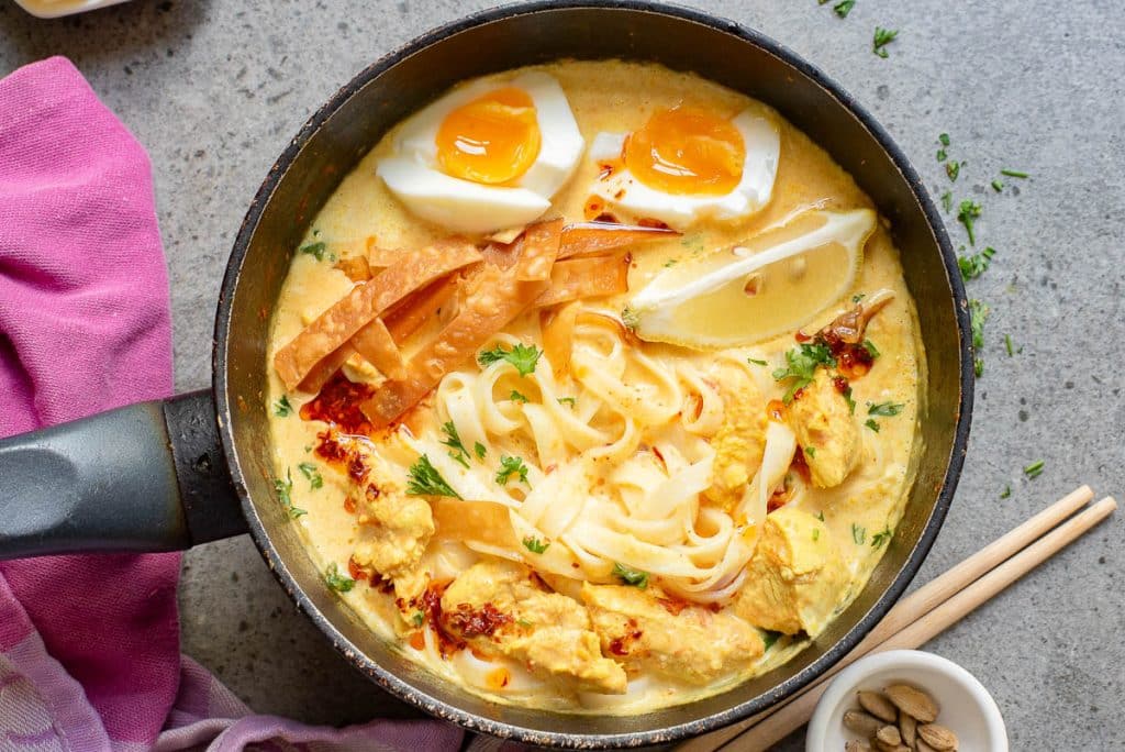 A bowl of creamy noodle soup with sliced chicken, soft-boiled eggs, and lemon wedges. Topped with crispy strips and herbs. Wooden chopsticks rest on the side. A pink cloth partially visible under the bowl.