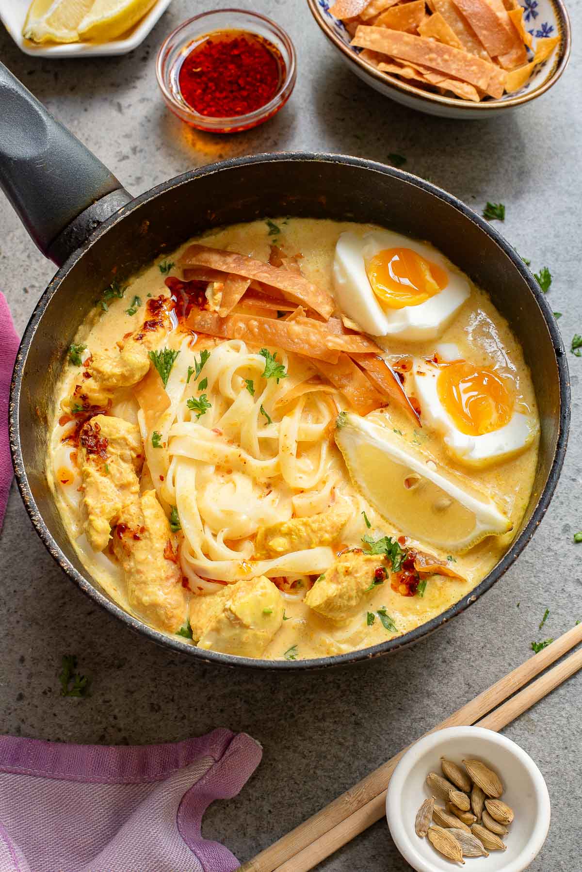 A pot of khao soi noodle soup with pieces of chicken, soft-boiled eggs, lemon wedges, and crispy wonton strips, garnished with fresh herbs. Surrounding the pot are small bowls of chili flakes, noodles, and cardamom pods. Chopsticks and a napkin are nearby.