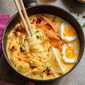A pan filled with creamy noodle soup topped with chicken pieces, soft-boiled eggs with runny yolks, crispy wonton strips, and a slice of lime. Chopsticks rest on the side. The dish is garnished with fresh herbs and chili oil.