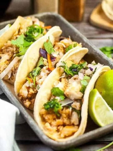 A tray of three tacos filled with chicken, cilantro, and onions. Lime wedges are on the side. A bunch of cilantro and a stack of tortillas are in the background. A gray cloth is partially visible on the left.