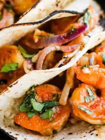 Three shrimp tacos filled with seasoned shrimp, sliced onions, and garnished with fresh cilantro are served on a black plate. A few lime wedges are placed next to the tacos. The tortillas are lightly charred.