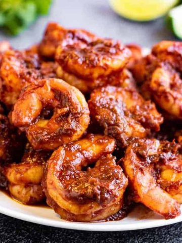 A pile of cooked shrimp covered in a rich, dark red sauce sits on a white plate. The shrimp are coated evenly with the sauce, appearing juicy and flavorful. A green garnish and lime wedges are blurred in the background.