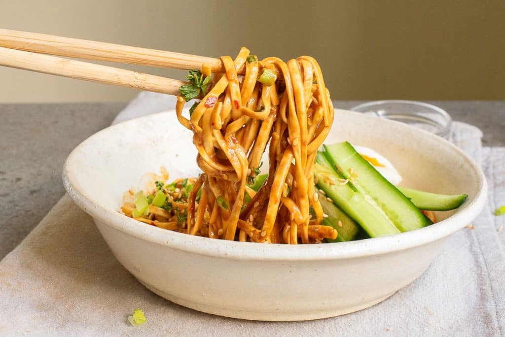 A bowl of noodles with a creamy sauce being lifted by chopsticks. Garnished with sliced cucumbers, sesame seeds, and green onions. Set on a textured gray surface.