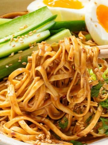 A bowl of noodles topped with sliced cucumbers, two halves of a soft-boiled egg, and sprinkled sesame seeds. The noodles are being lifted with chopsticks and garnished with fresh herbs.
