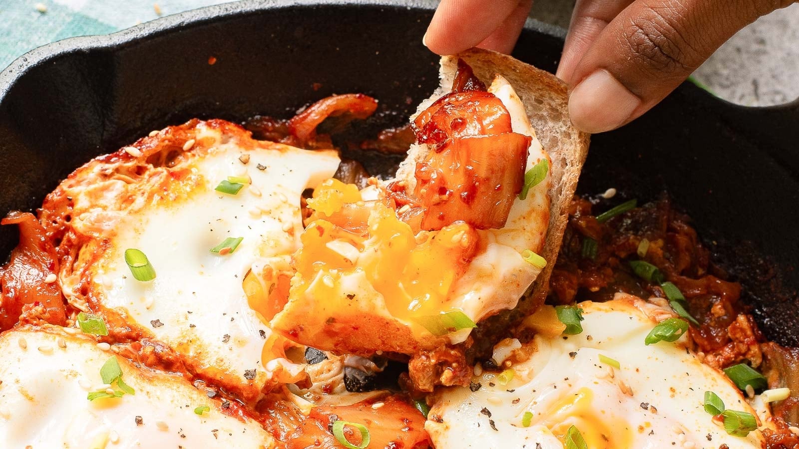 A hand dips bread into a skillet of shakshuka, featuring poached eggs, tomato sauce, and garnished with chopped green onions.