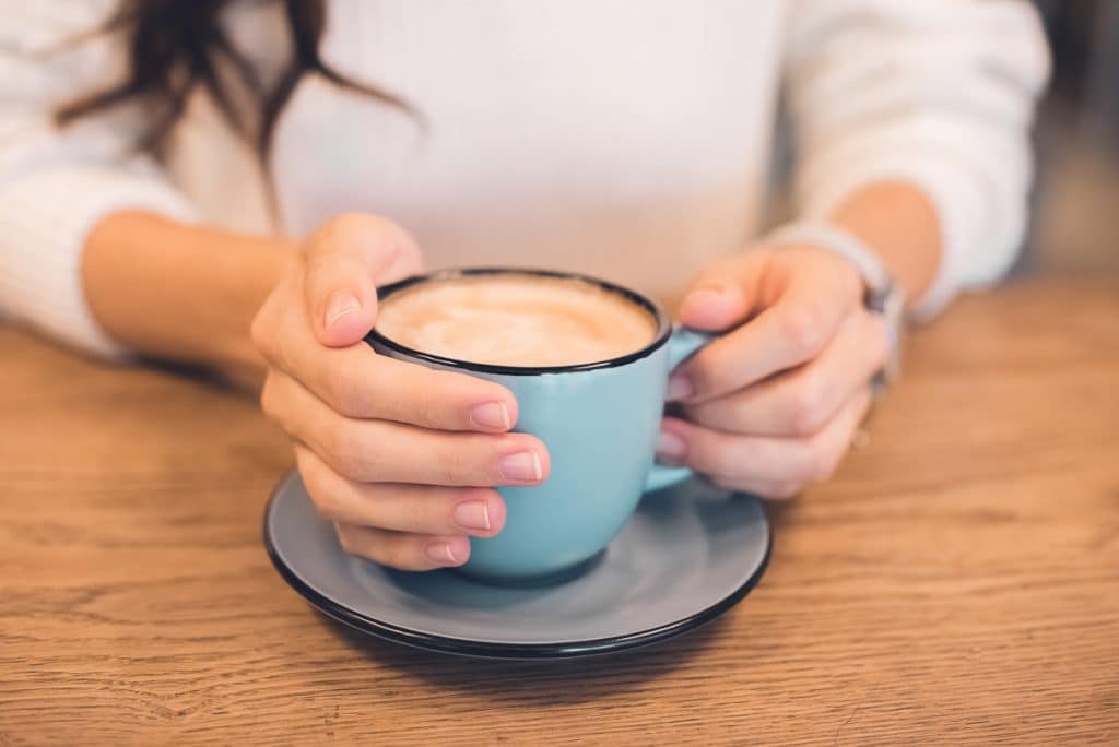 A person in a white sweater holds a blue cup filled with frothy coffee, resting on a matching saucer atop a wooden table. The hands are gently cradling the cup.