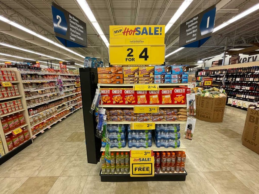 Supermarket aisle with a sale display showcasing various snacks, including Cheez-It boxes, bottled water, and beverages. A large sign on top reads "MVP Hot Sale! 2 for $4" and lists prices. Aisles 1 and 2 are visible with more products.