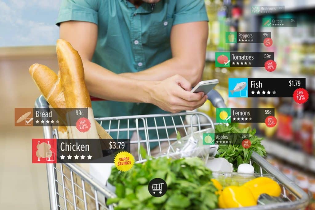 A person checks their phone while leaning on a shopping cart filled with groceries like bread, lettuce, and lemons. Floating icons show product names, prices, discounts, and ratings for items like fish, tomatoes, and chicken in a grocery store aisle.