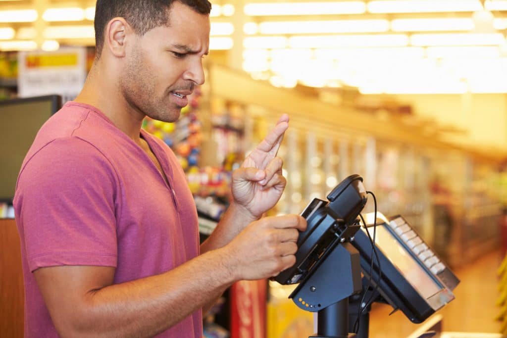 Man in a pink shirt using a payment terminal at a store checkout. He appears anxious and is crossing his fingers on one hand while pressing buttons on the terminal with the other. Blurred store shelves are visible in the background.