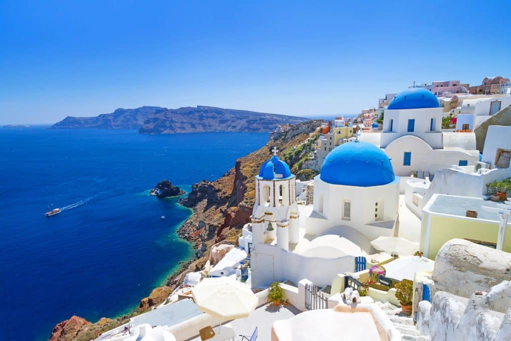 White buildings with bright blue domes overlook the deep blue Aegean Sea on a sunny day in Santorini, Greece, with rugged cliffs and a boat in the water in the background.