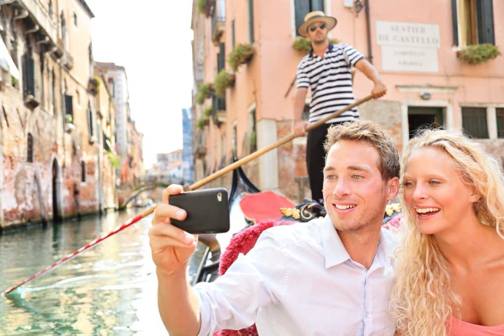 A smiling couple takes a selfie while riding a gondola in a Venetian canal, with a gondolier in a striped shirt standing behind them and historic buildings lining the water.