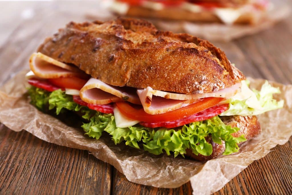 A rustic sandwich on a brown paper with layers of lettuce, sliced tomatoes, salami, cheese, and ham, all inside a crusty whole-grain roll. The sandwich is on a wooden surface with a blurred sandwich in the background.