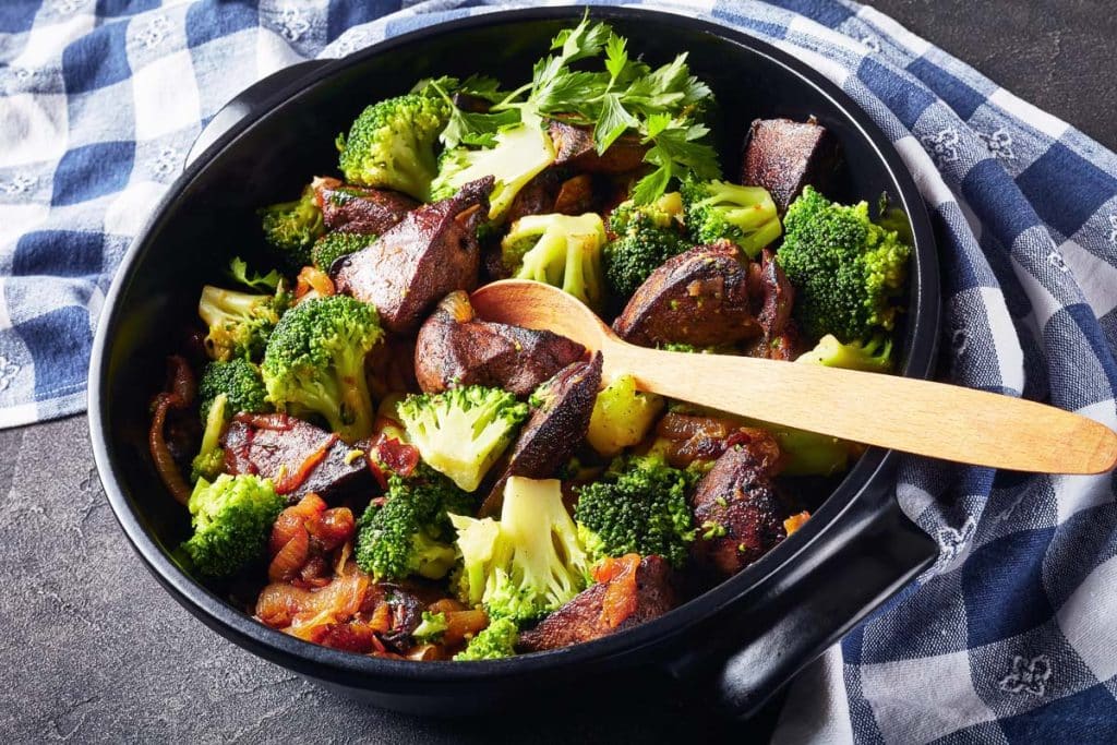 A black skillet filled with roasted broccoli florets and slices of beef, garnished with fresh parsley, sits on a checkered blue and white tablecloth. A wooden spatula rests on the skillet.