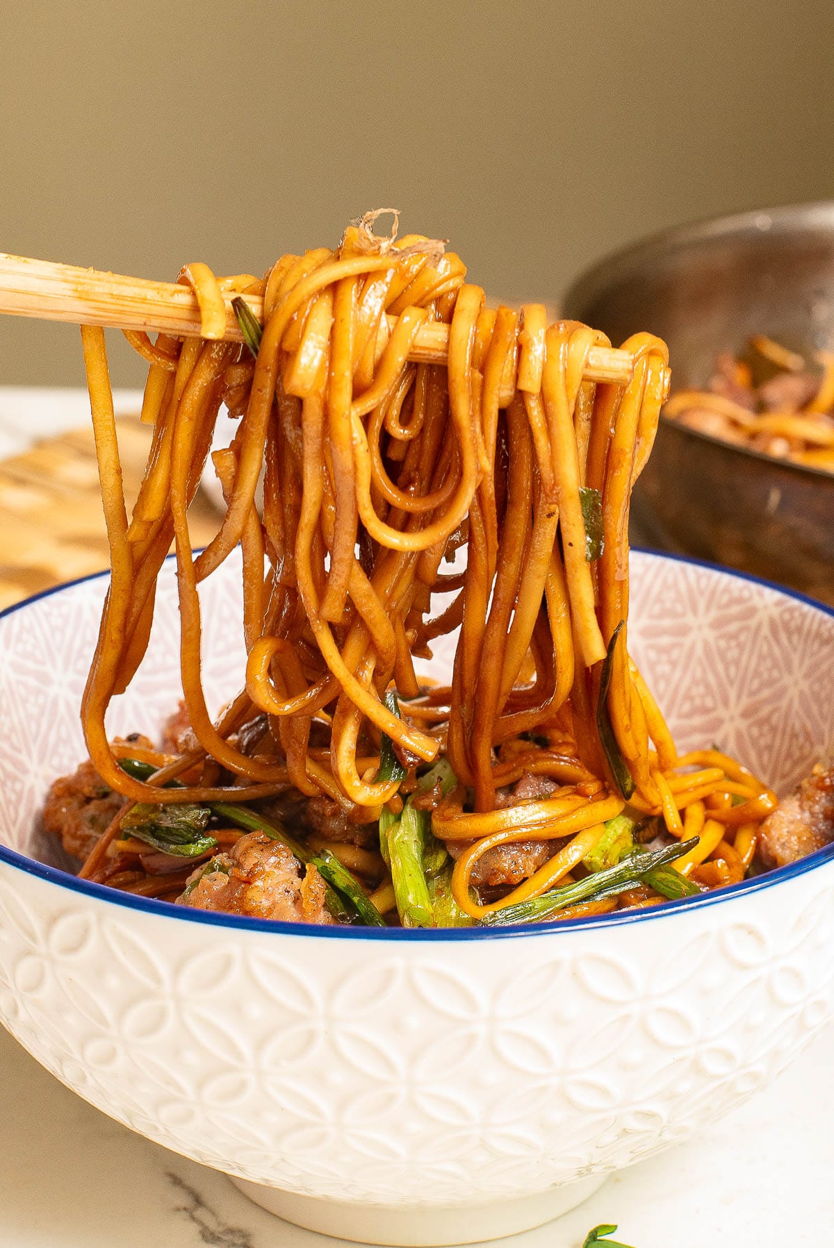 A bowl of saucy ramen noodles with greens and meatballs. Chopsticks are holding up a portion of the noodles above the bowl. The dish is in a patterned white bowl on a marble countertop.