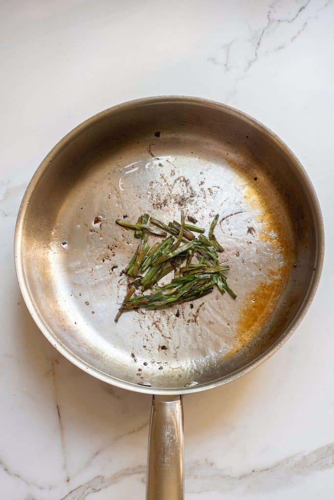 A stainless steel frying pan with remnants of cooked green herbs, possibly rosemary, resting in the center. The pan shows signs of being used, with oil and seasoning residue visible. It sits on a white surface.