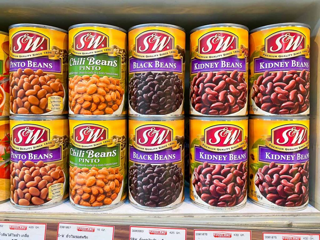 Cans of S&W beans on a store shelf, featuring pinto chili beans, black beans, and kidney beans. Each can has a yellow label with images of the beans.