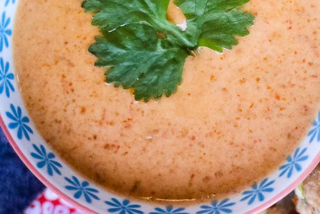 A close-up of a creamy orange soup garnished with a sprig of fresh green cilantro. The soup is served in a white bowl with a blue floral pattern along the rim. A red and white cloth is partially visible beneath the bowl.