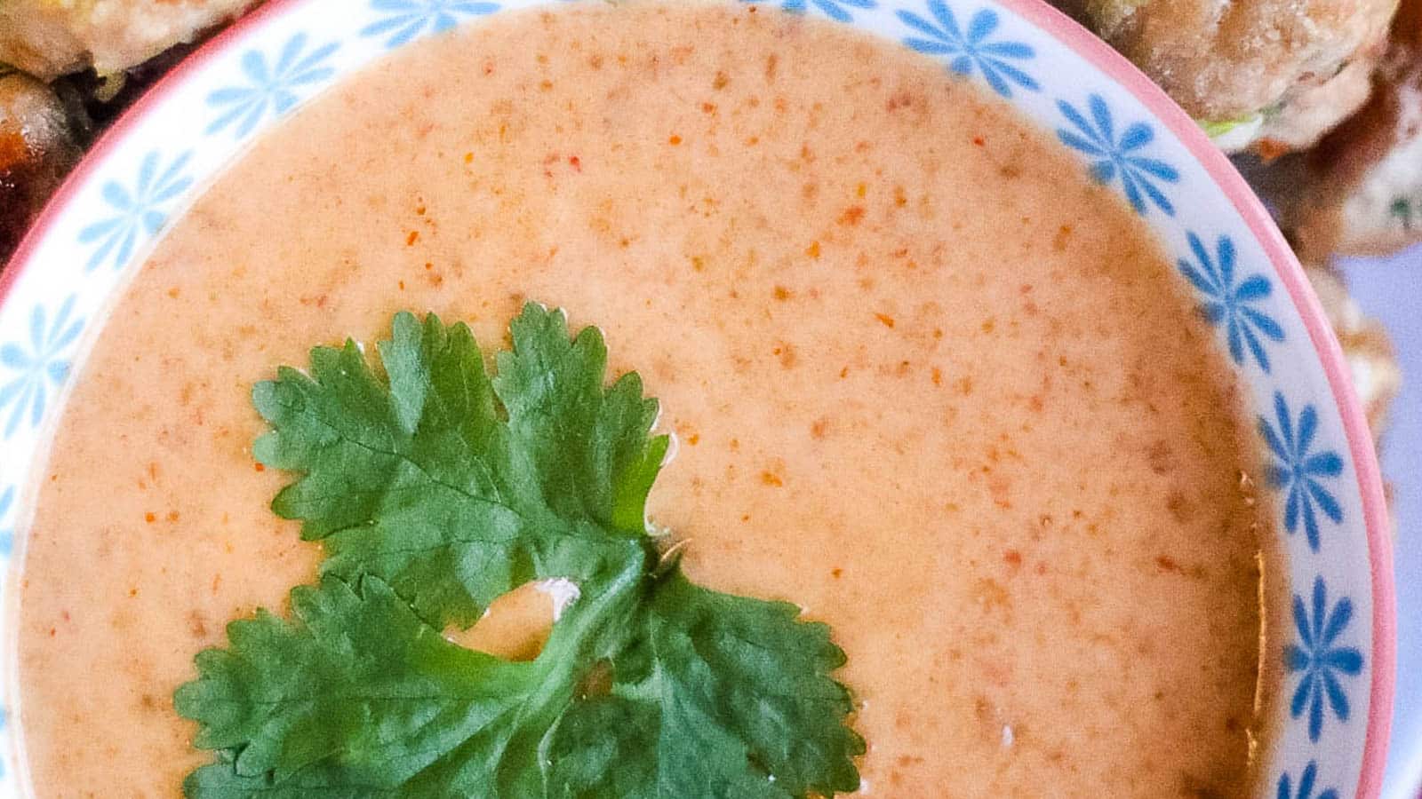 Close-up of a small bowl filled with a creamy, orange-hued sauce garnished with a fresh cilantro leaf. The bowl has a white interior with a light blue floral rim.