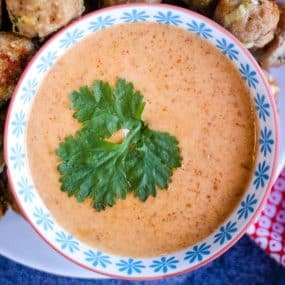 A close-up of a bowl filled with creamy orange-colored sauce, garnished with fresh cilantro leaves. The bowl has a blue patterned rim and is placed on a plate, surrounded by partially visible food items in the background.