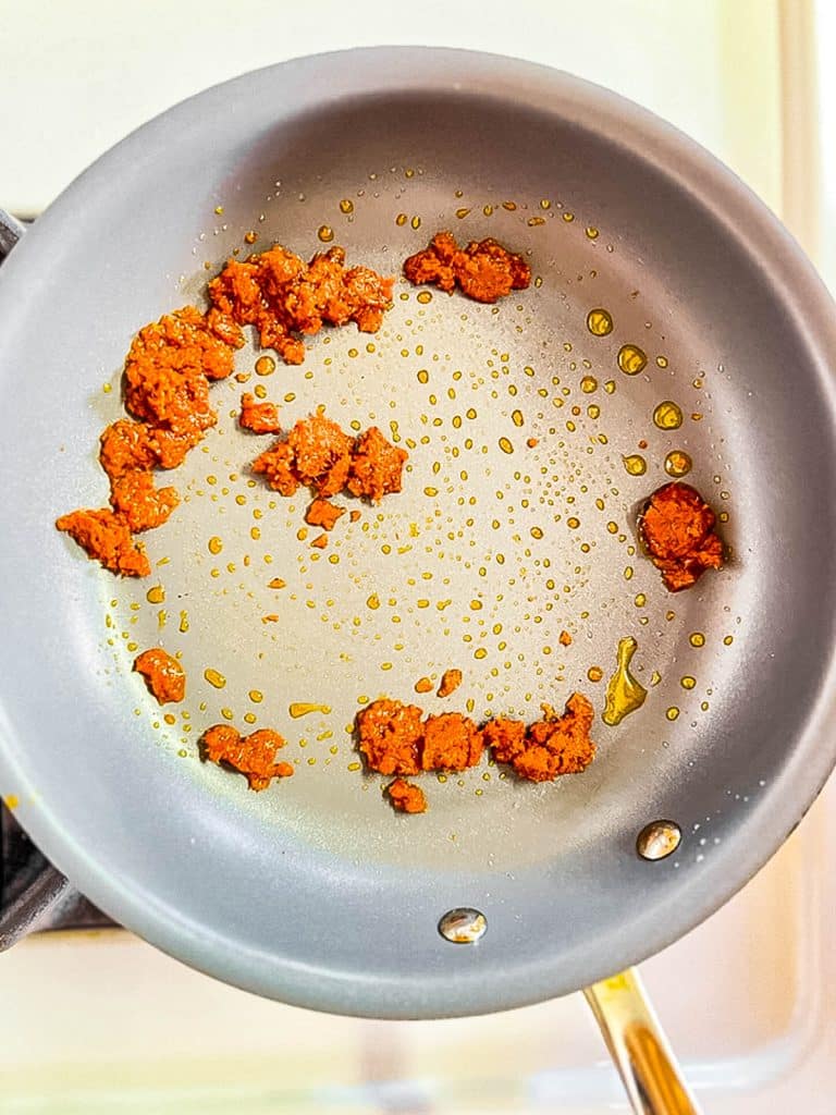 Ground chorizo being cooked in a gray non-stick frying pan. The chorizo pieces are spread in a circle, surrounded by small droplets of oil. The pan is on a stove with a light background.