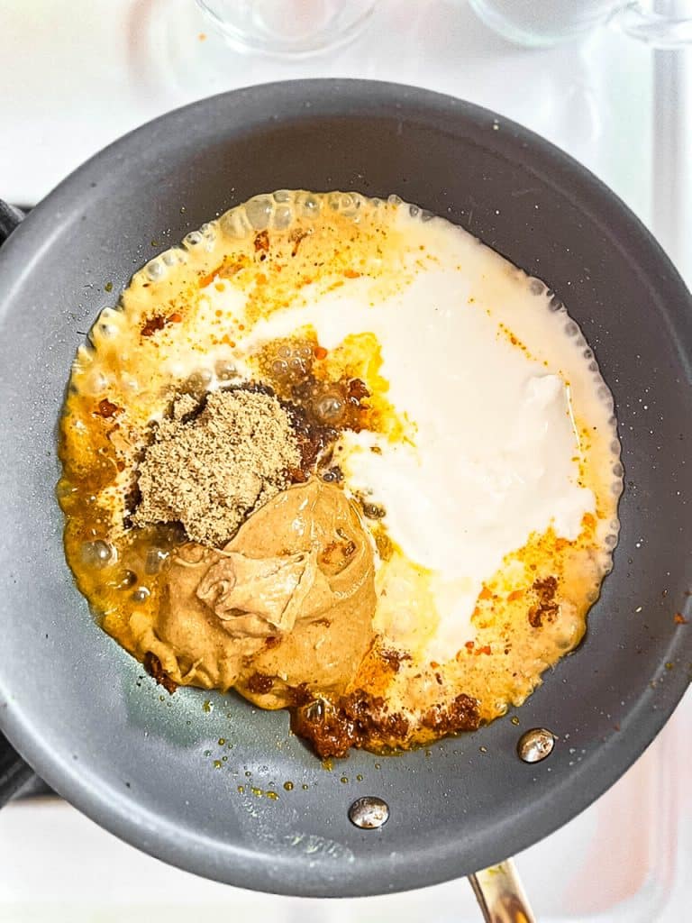A frying pan contains cream, a dollop of peanut butter, a mound of brown sugar, and spices, starting to mix together. The ingredients are beginning to bubble and blend, suggesting the preparation of a creamy dish or sauce.