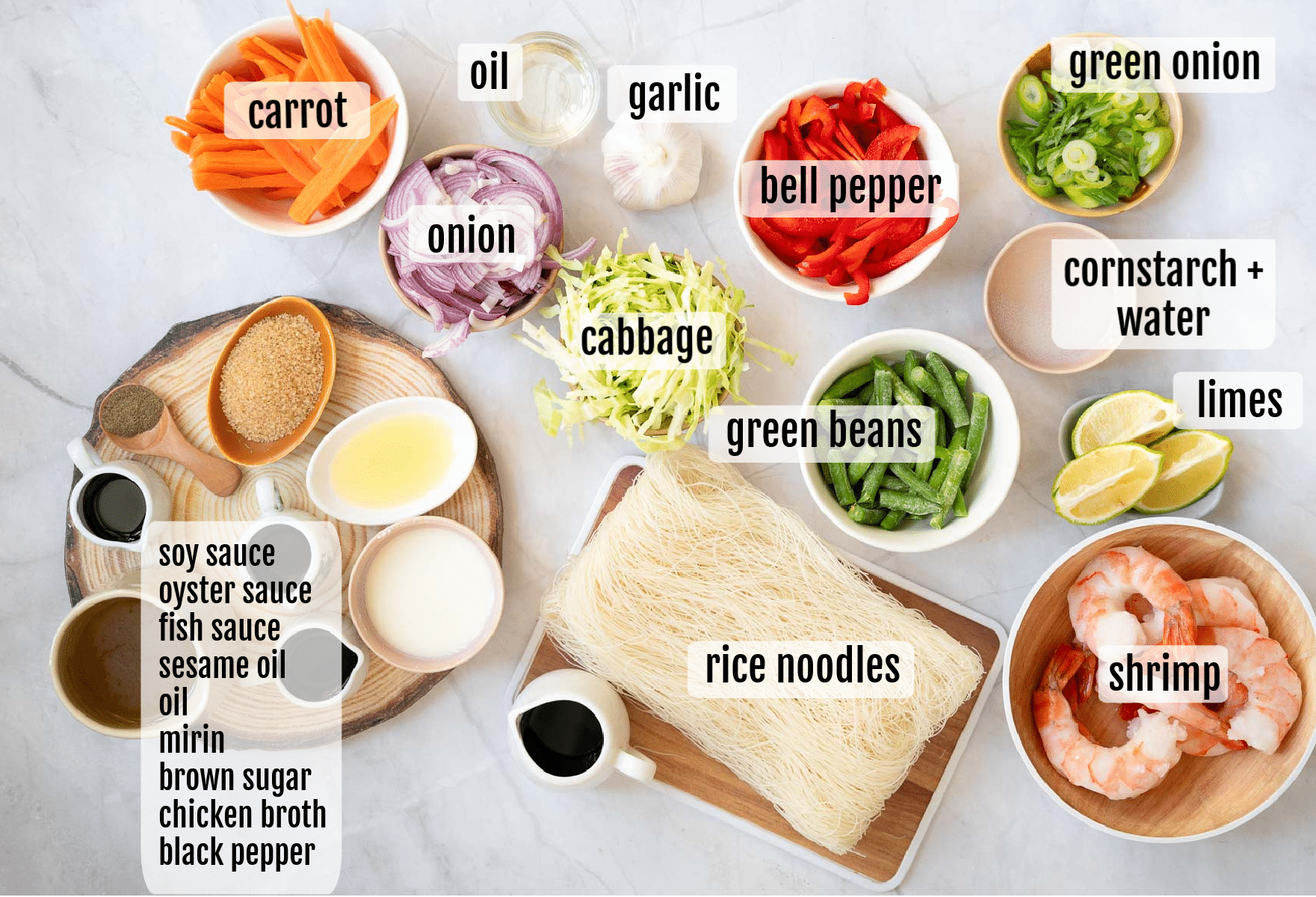 A top-down view of ingredients for a noodle dish, including rice noodles, shrimp, cabbage, onion, carrot, bell pepper, green beans, green onion, limes, garlic, oil, cornstarch with water, and various sauces and spices.