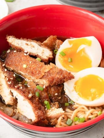 A red bowl filled with ramen noodles, topped with crispy breaded chicken slices, two halves of a soft-boiled egg, chopped green onions, and sesame seeds. Chopsticks and a small dish of green onions are beside the bowl.