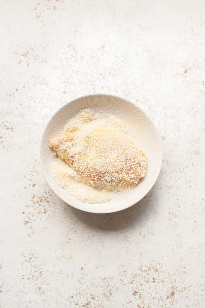 A piece of raw chicken coated in breadcrumbs sits on a white plate, with extra breadcrumbs scattered around the plate on a light, textured surface.