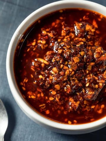 A white bowl filled with chili oil containing visible chili flakes, sesame seeds, and bits of garlic, placed on a dark gray surface.