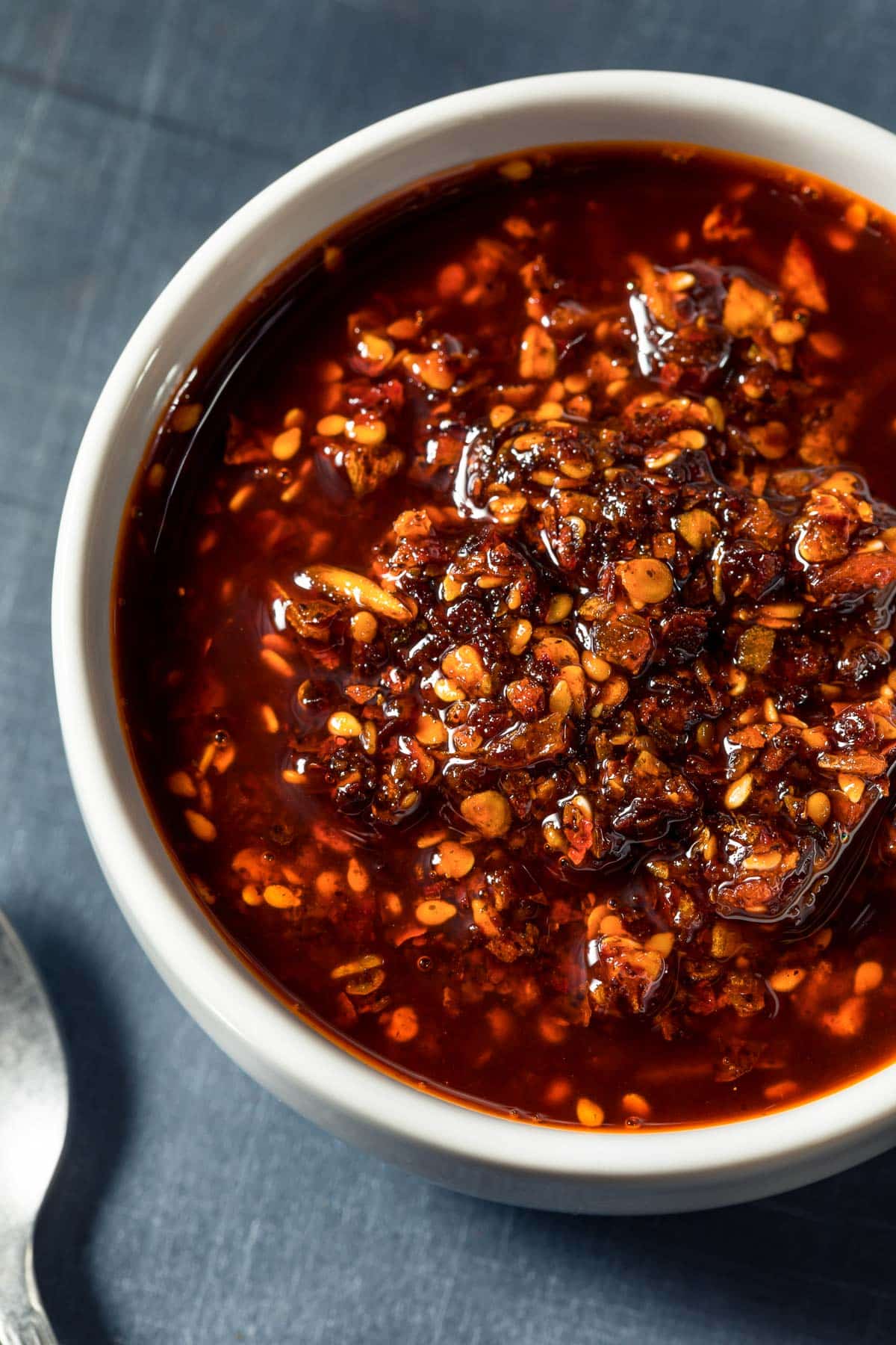 A white bowl filled with chili oil containing visible chili flakes, sesame seeds, and bits of garlic, placed on a dark gray surface.