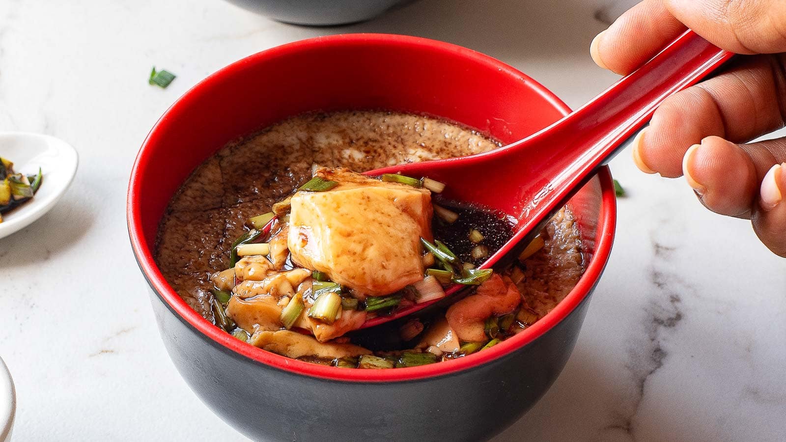 A hand holds a red spoon lifting a piece of tofu from a red bowl filled with soup, garnished with chopped herbs and sauce, reminiscent of a comforting Chinese Steamed Egg recipe.