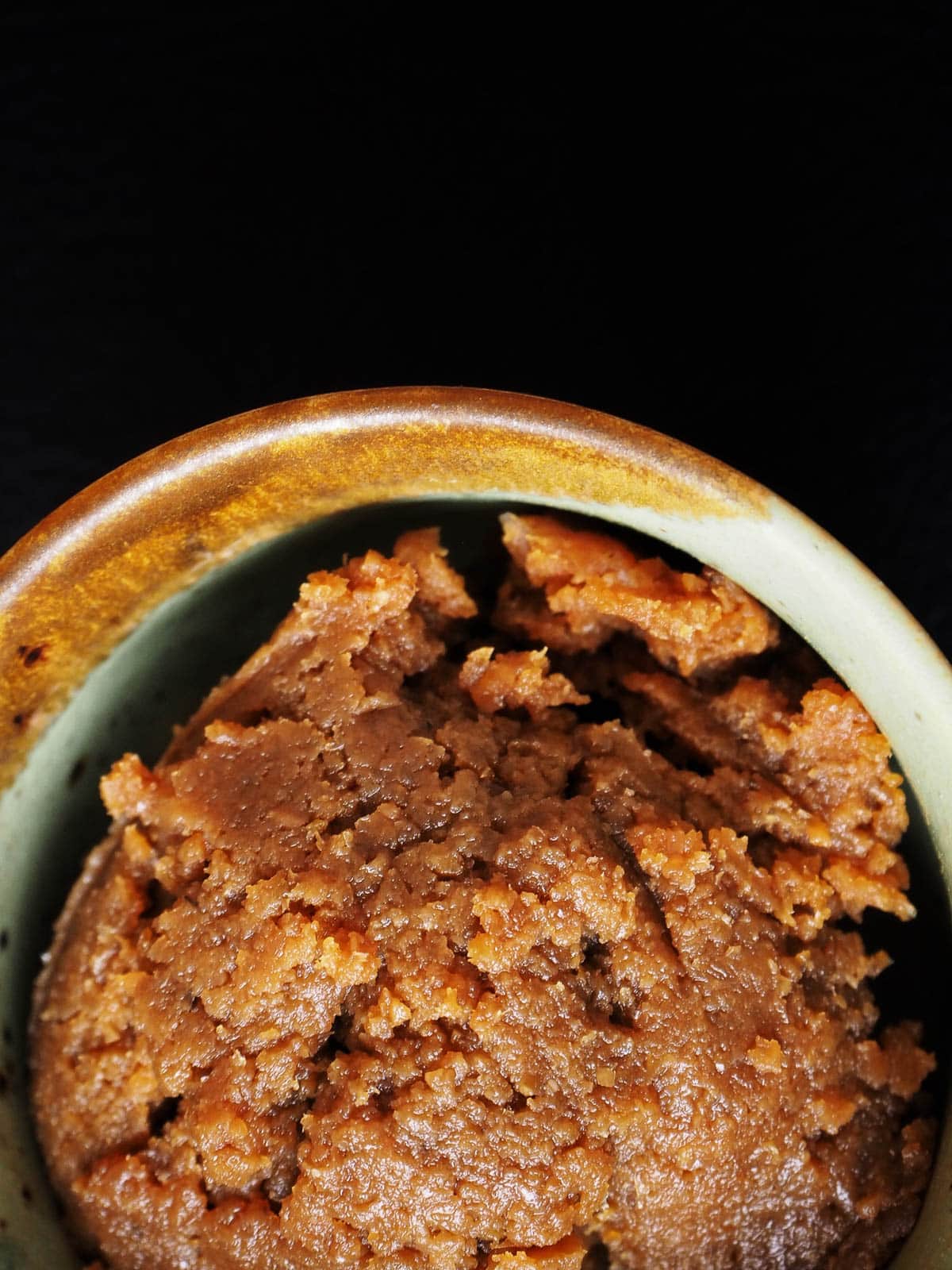 A close-up of a bowl filled with brown miso paste, showing its coarse, chunky texture against a dark background. The bowl has a brown and greenish rim.