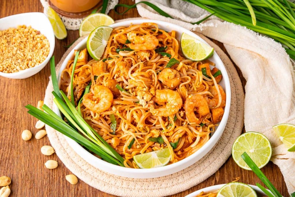 A white bowl of shrimp pad thai with tofu, crushed peanuts, green onions, and lime wedges sits on a woven mat, surrounded by fresh green onions, peanuts, and lime slices on a wooden table.