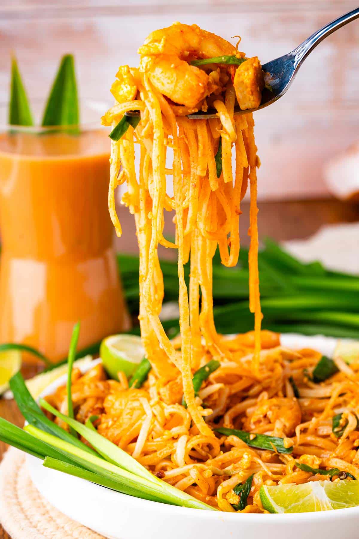 A fork lifts a serving of Pad Thai noodles with shrimp and vegetables from a white plate, garnished with lime wedges and green onions; a glass of Thai iced tea is in the background.
