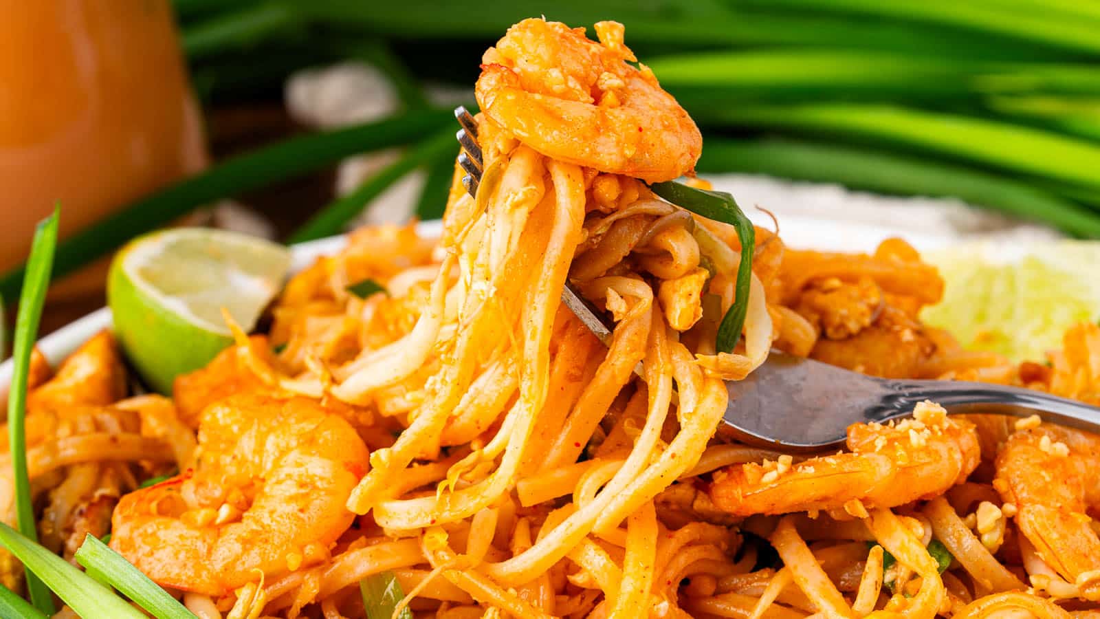 A close-up of shrimp pad thai, with noodles, shrimp, green onions, and a fork lifting a bite. A lime wedge and blurred green onions are visible in the background.