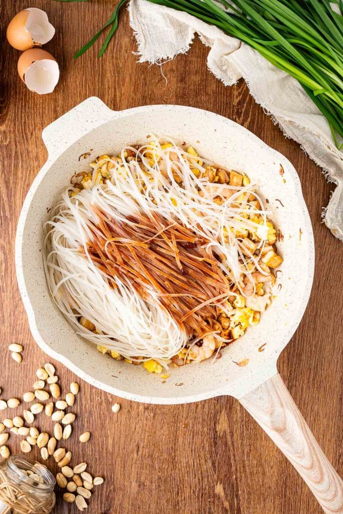 A frying pan on a wooden surface contains rice noodles, sauce, eggs, peanuts, and shrimp. Chopped green onions, whole peanuts, garlic, and cracked eggshells are scattered nearby.