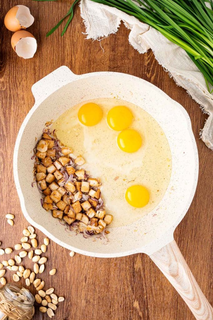 A white skillet on a wooden surface contains three raw eggs and sautéed diced onions. Around the skillet are eggshells, chives, a linen cloth, peanuts, and a small bowl of peanuts.