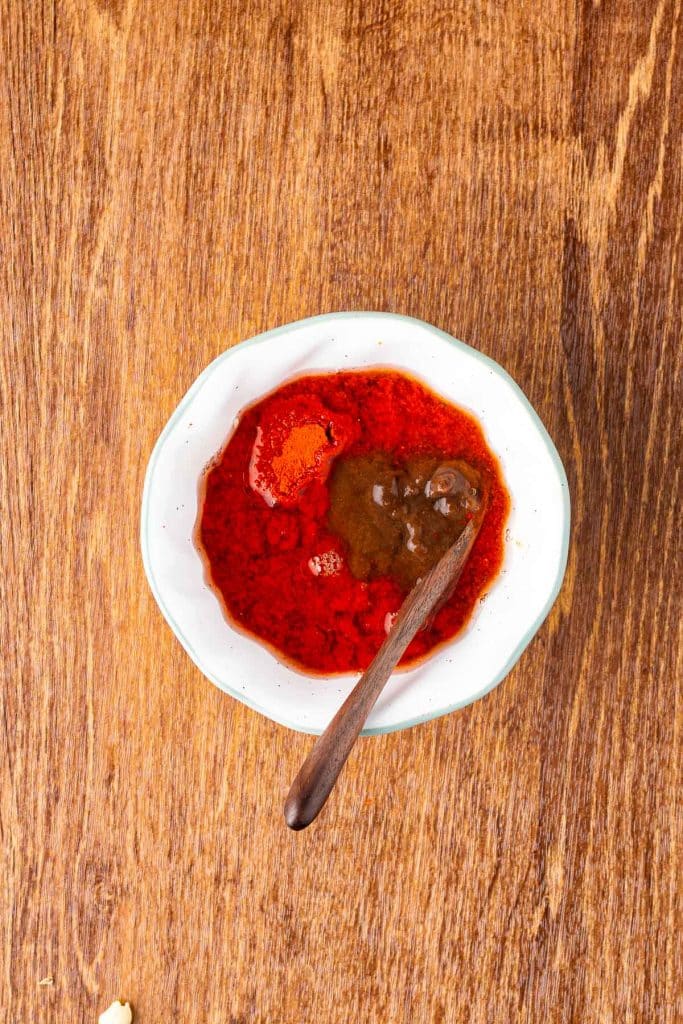 A small white bowl filled with red liquid and spices sits on a wooden surface. A wooden spoon rests inside the bowl.