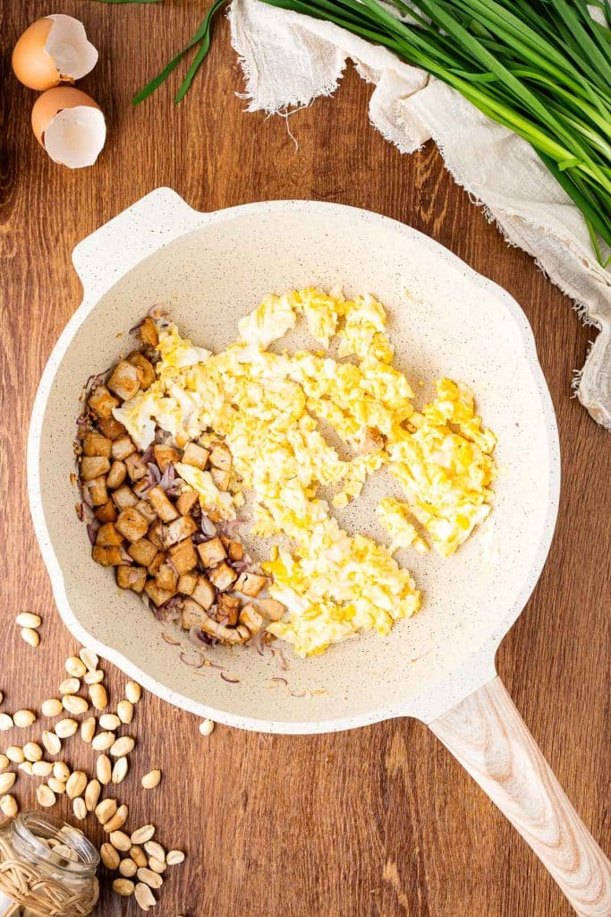 A frying pan on a wooden surface contains scrambled eggs and diced cooked mushrooms. Nearby are scattered peanuts, eggshells, green onions, and a piece of cloth.