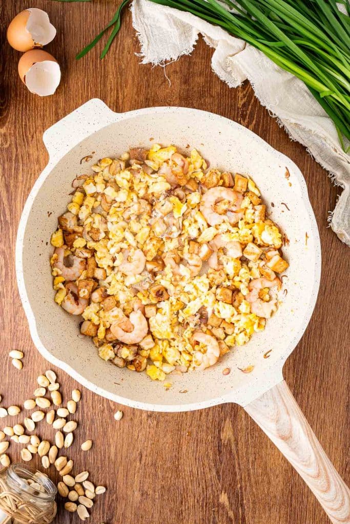 A white skillet filled with scrambled eggs, shrimp, and diced tofu sits on a wooden surface, surrounded by fresh chives, two cracked eggs, pistachios, and a linen cloth.