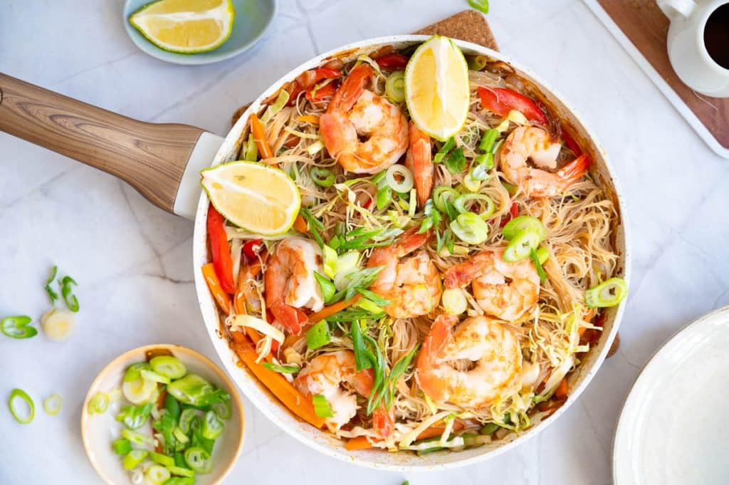 A pan filled with shrimp, sliced bell peppers, cabbage, and rice noodles, garnished with green onions and lemon wedges, sits on a marble surface. A small bowl of sliced green onions and a lemon wedge are nearby.